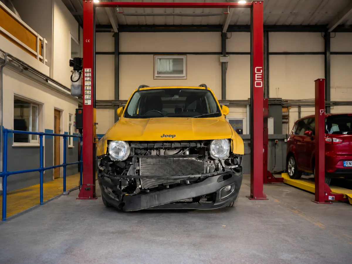 Yellow Jeep Renegade with front-end damage raised on a two-post ramp in a UK salvage workshop