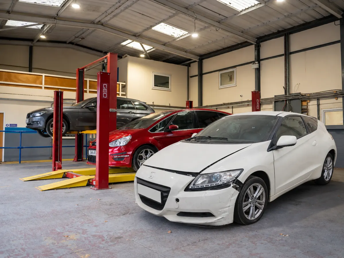 White Honda CR-Z coupé in a workshop alongside other vehicles awaiting assessment, with only minor cosmetic damage visible