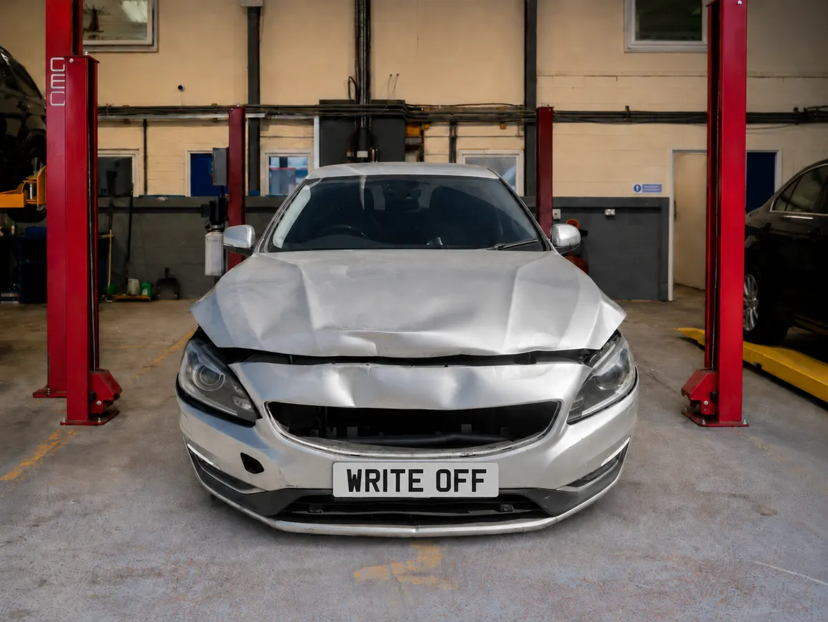 Silver Volvo S60 in a workshop with a crumpled bonnet and collapsed grille from a front-end impact, sitting between cosmetic damage and structural failure