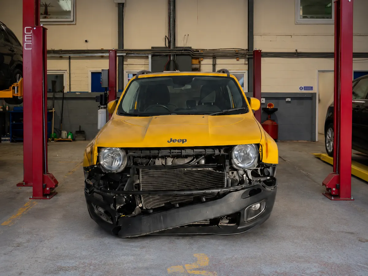 Yellow Jeep Renegade raised on a workshop ramp with its front bumper removed, exposing the radiator and front-end damage
