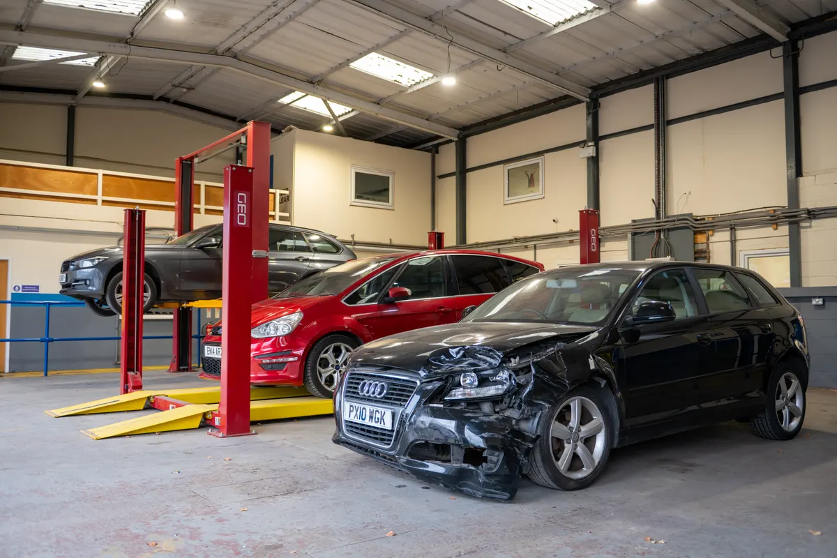 Vehicles in a UK salvage workshop, including a black Audi with front-end damage and a car raised on a two-post ramp